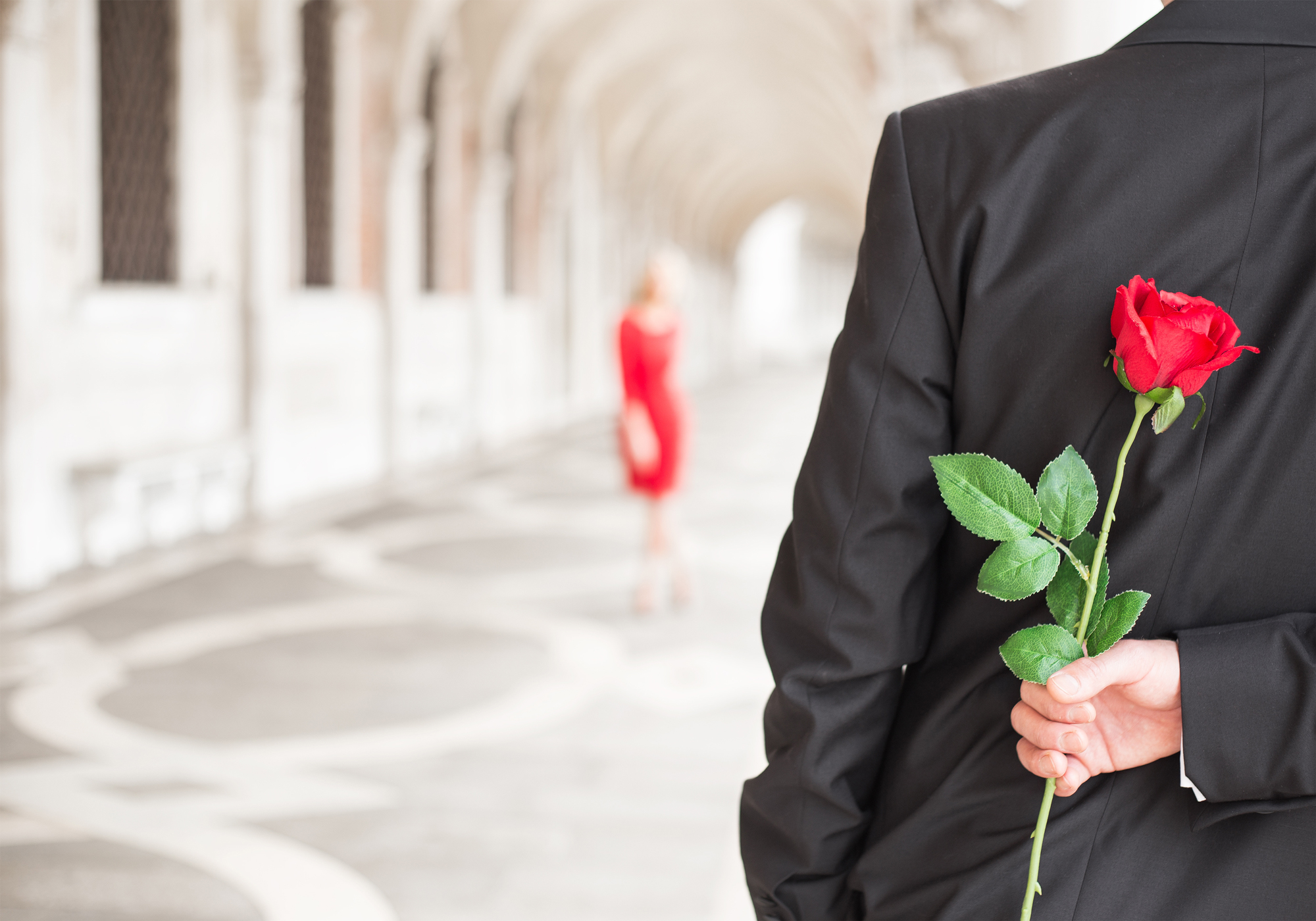 Man holding red rose behind back as woman in red dress approaches